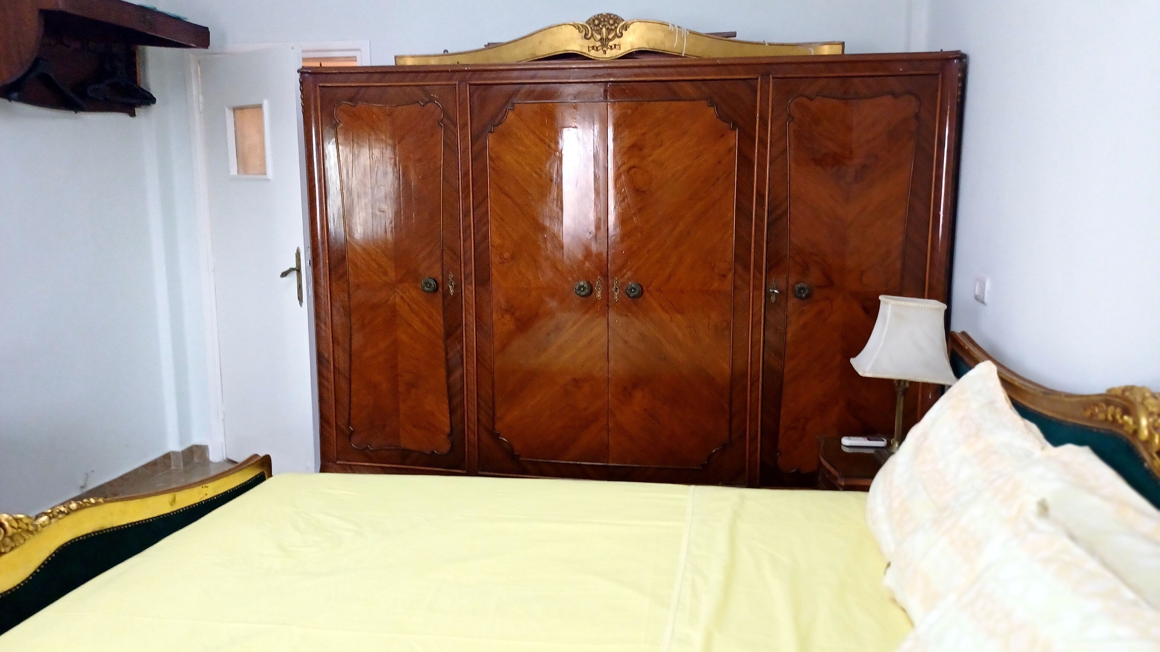 A bedroom with a large wooden wardrobe, a bed with a yellow bedspread, and a bedside table with a lamp.