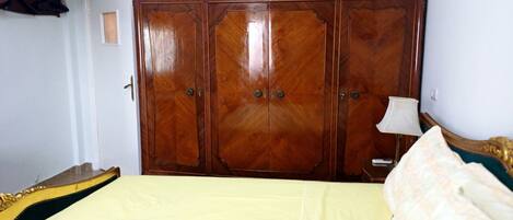 A bedroom with a large wooden wardrobe, a bed with a yellow bedspread, and a bedside table with a lamp.