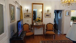 A room with a television, two chairs, a table with a vase of flowers, a large mirror, and a chandelier. - Elegant 2-Bedroom Apt with City View in Agouza. (Giza Governorate)