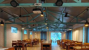 A spacious indoor dining area with high ceilings, exposed beams, and a mix of brick and wooden walls. Tables and chairs are arranged for dining. - Kumaoni Karavan Camps Resort (Nainital)