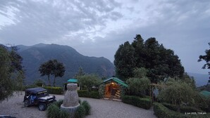 A rustic lodge with a stone centerpiece, surrounded by lush greenery and a mountain backdrop. - Kumaoni Karavan Camps Resort (Nainital)