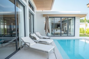 Un área moderna junto a la piscina con sillas de descanso, una sala con paredes de vidrio y vista despejada al agua.