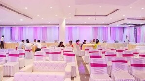 Uma sala de banquetes com fileiras de cadeiras brancas, cada uma com uma fita rosa, e um palco na frente.