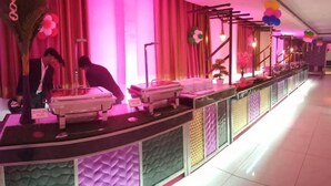 Uma área de buffet com esquema de cores rosa e roxo, apresentando um balcão com displays de comida e uma planta decorativa.