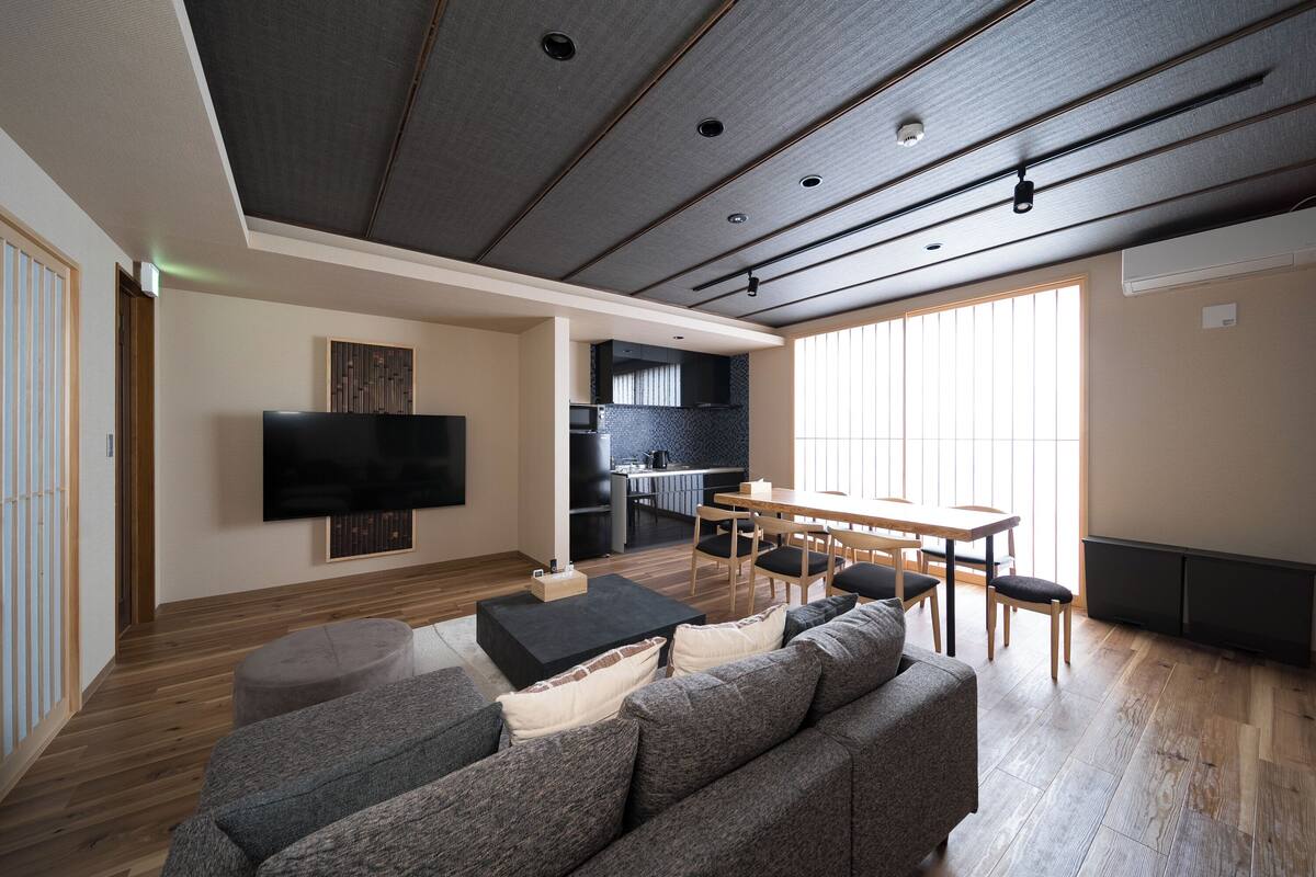 A modern living room with a flat-screen TV, grey sofa, wooden flooring, and a dining area with a table and chairs.