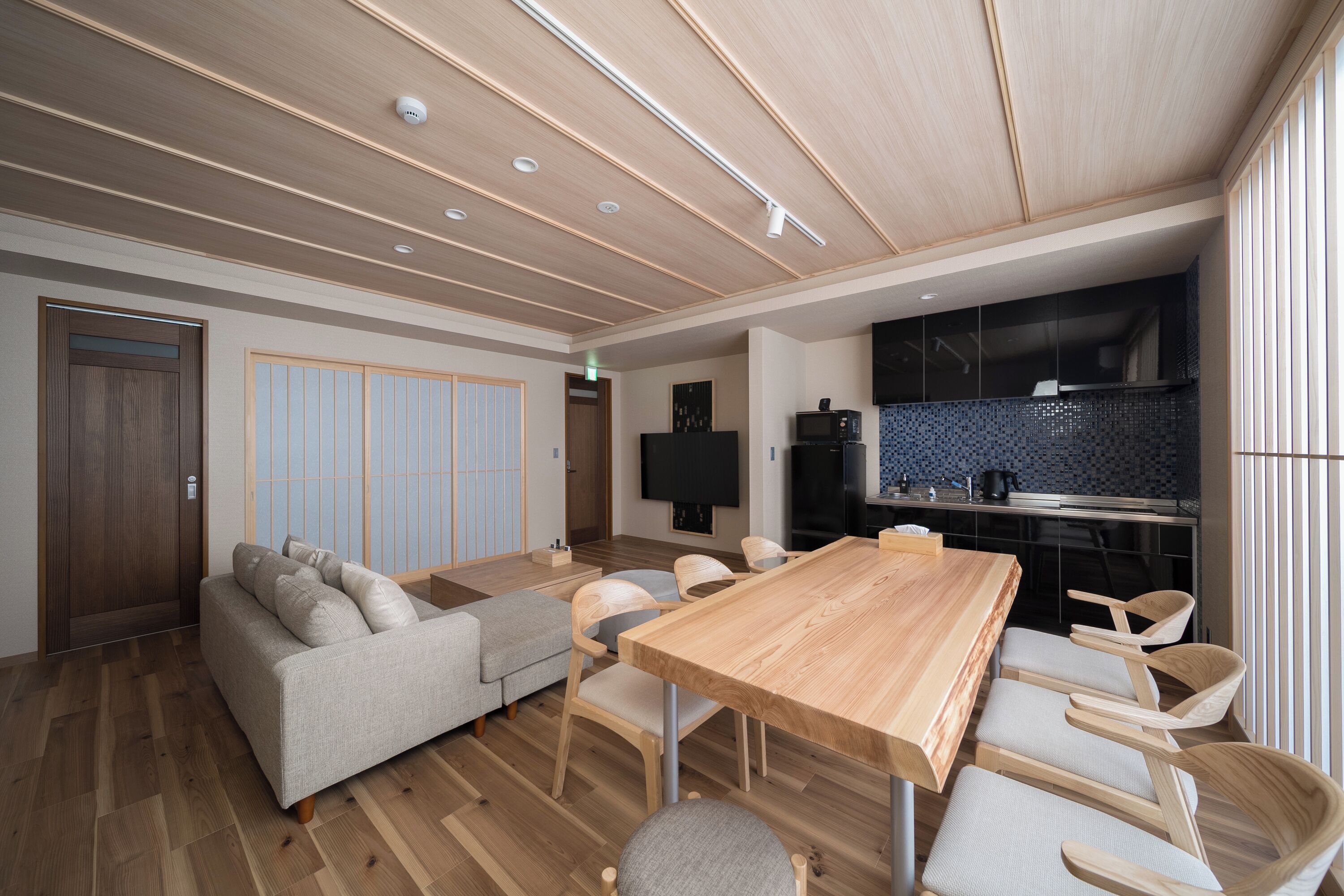 A modern living room with a wooden ceiling, a sofa, a dining table with chairs, and a kitchen area with a TV and appliances.