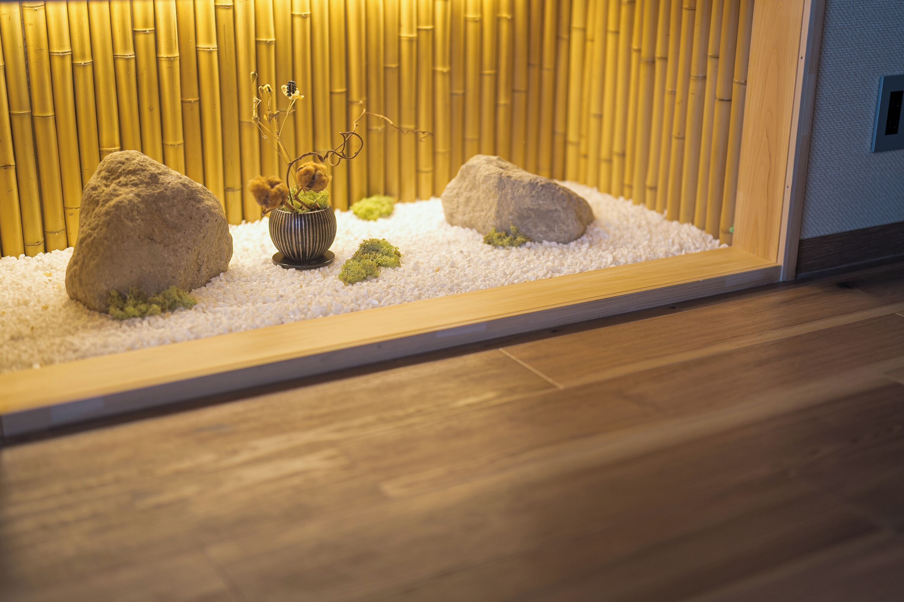 A room with a wooden floor, a bamboo wall, and a small indoor garden with rocks and a plant in a bowl.