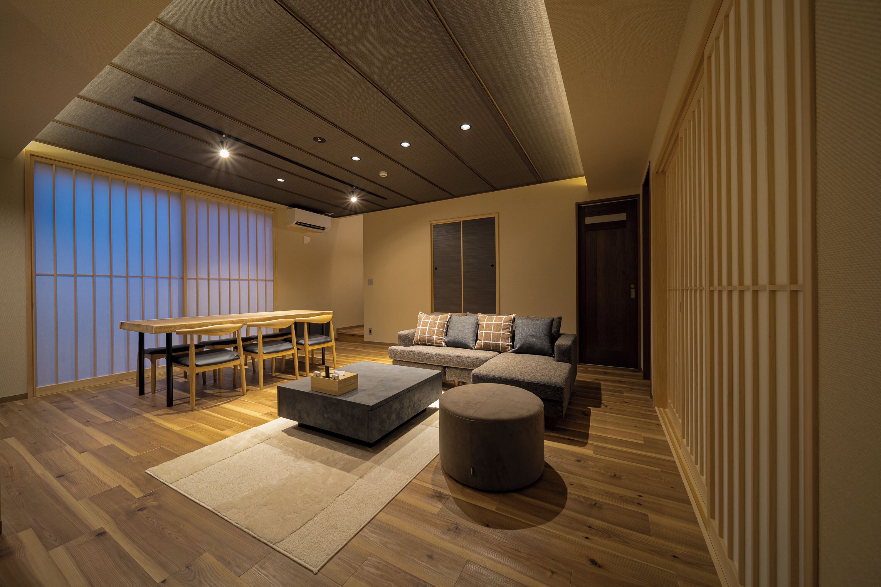 A modern living room with a wooden floor, a sofa, a dining table with chairs, and a large window with blinds.