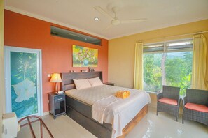 A bedroom with a bed, nightstand, lamp, two chairs, a ceiling fan, a window with curtains, and a painting on the wall. - Maria Secret Villa (Sukasada)