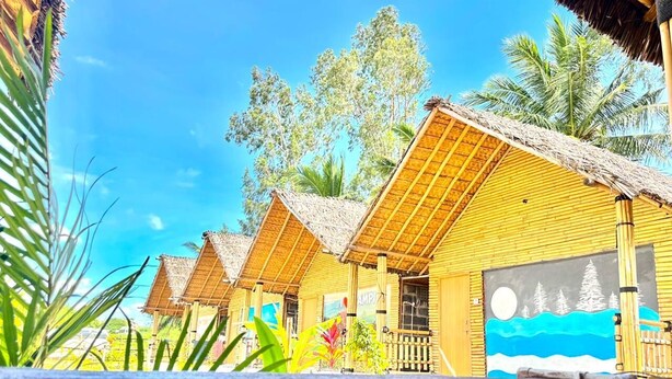 A row of thatched-roof cottages with a mural on one side, surrounded by palm trees and clear skies. - Bliss Hampi Eco Resort (Koppal)