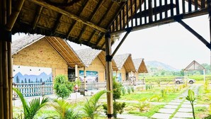 A covered outdoor area with thatched-roof structures, a stone pathway, and lush greenery. - Bliss Hampi Eco Resort (Koppal)