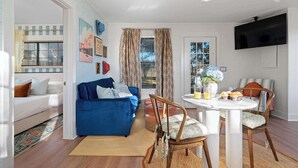 Una sala de estar con un sofá azul, una mesa de comedor con sillas, un televisor montado en la pared y una ventana con cortinas.