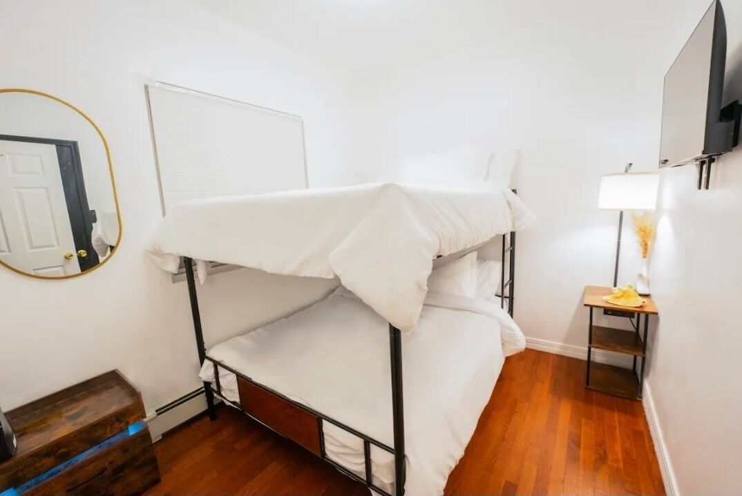 A bunk bed room with a wooden floor, a small table with a lamp, and a mirror on the wall.