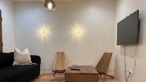 A modern living room with a black sofa, wooden chairs, a coffee table, a TV mounted on the wall, and two wall-mounted lights. - Marvelous 3-bedroom house with AC in Inglewood (Inglewood)