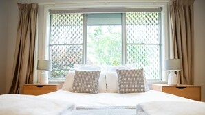 Een net opgemaakt tweepersoonsbed met twee kussens, een nachtkastje met een lamp en een raam met een gestreept raambekleding.