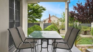 Een overdekt terras met een tafel en vier stoelen, een uitzicht op een woonwijk en een omheinde tuin.