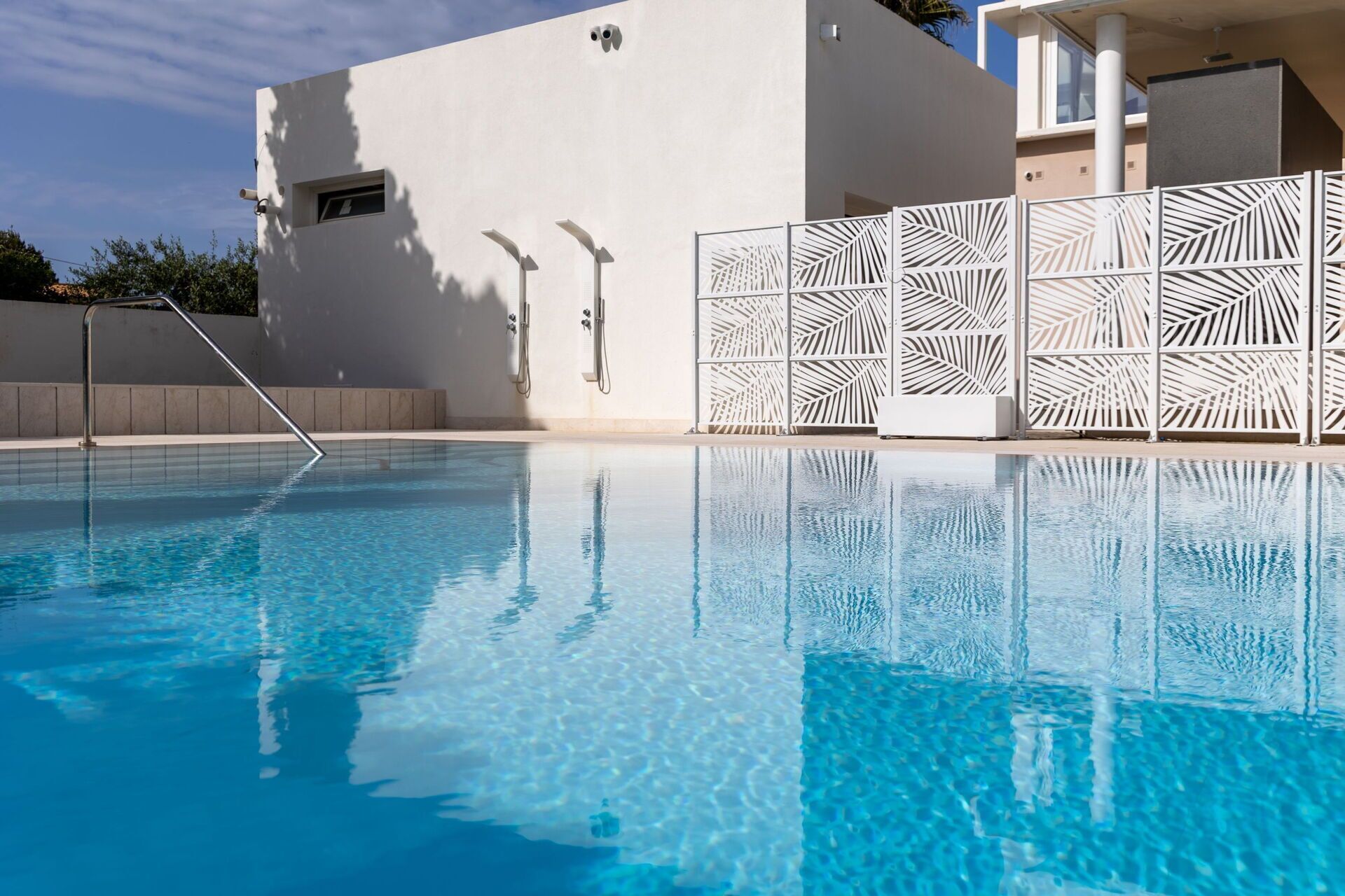 A clear swimming pool with a modern design, a white wall with a small window, and a decorative white fence with leaf patterns.