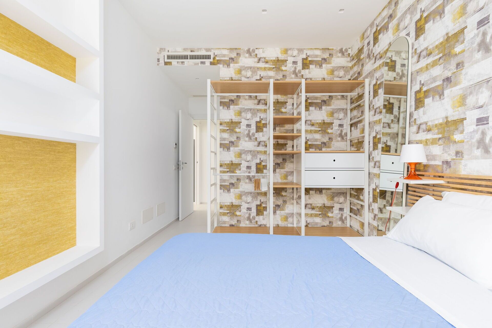A bedroom with a bed, a wardrobe with multiple shelves, a bedside table with a lamp, and a window.