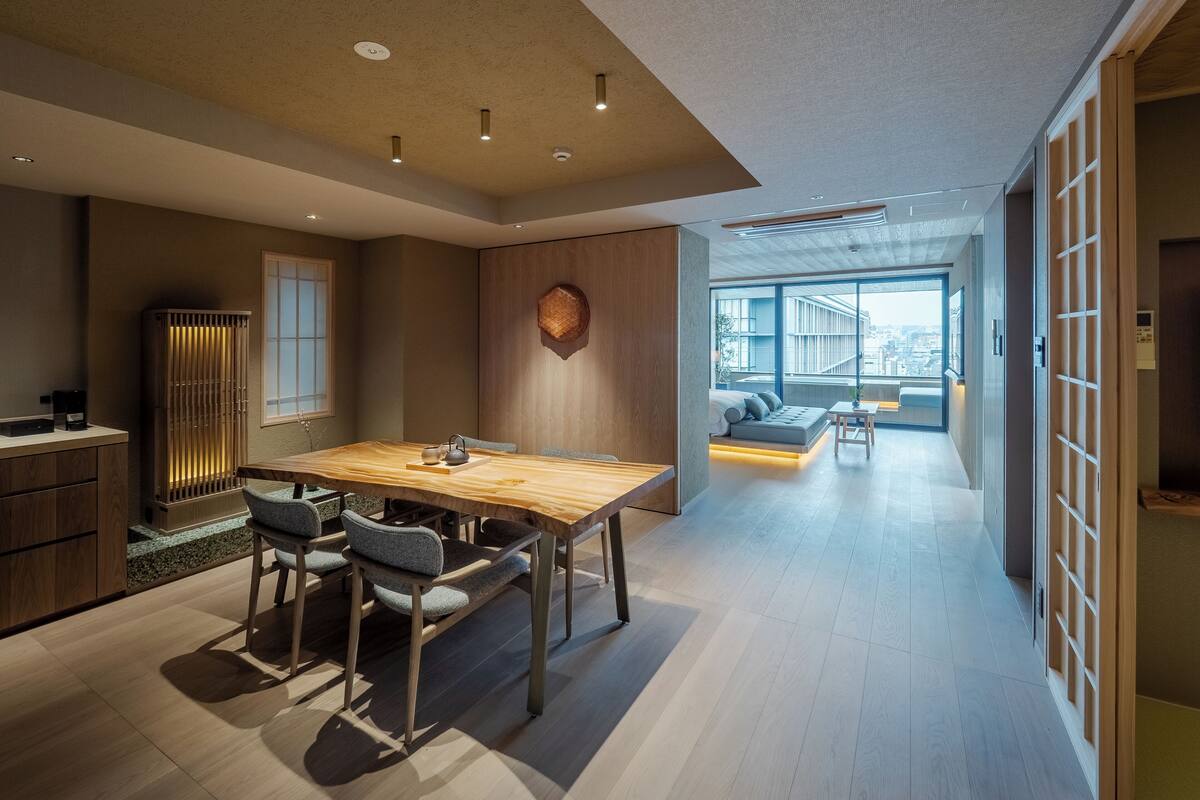 A modern interior with a wooden dining table, grey upholstered chairs, a built-in kitchen, and a view to the outside.