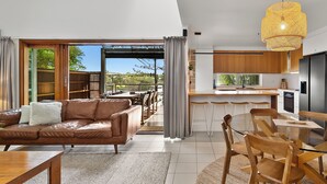 A modern kitchen with a built-in oven, a dining area with a glass table and wooden chairs, a leather sofa with a throw pillow, and a large window overlooking a garden.