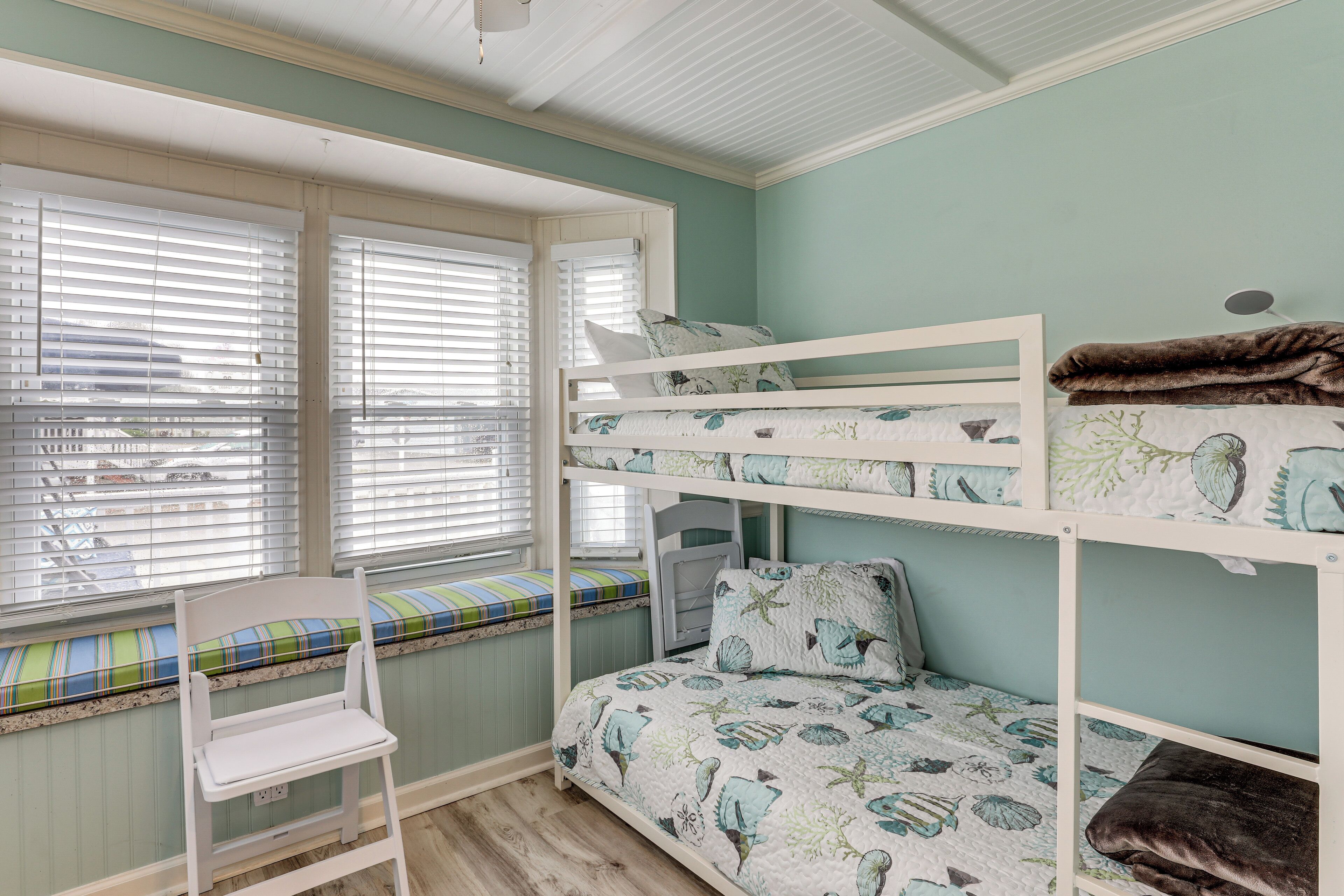 Ein Stockbett mit gemustertem Bettzeug, eine Fensterbank mit gestreifter Sitzkissen und ein weißer Stuhl.
