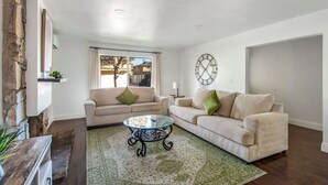 Un salón con dos sofás, una mesa de centro de vidrio, un reloj decorativo y una ventana con cortinas.