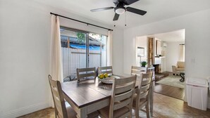 Un comedor con una mesa y sillas de madera, un ventilador de techo y vista al patio trasero a través de una ventana.
