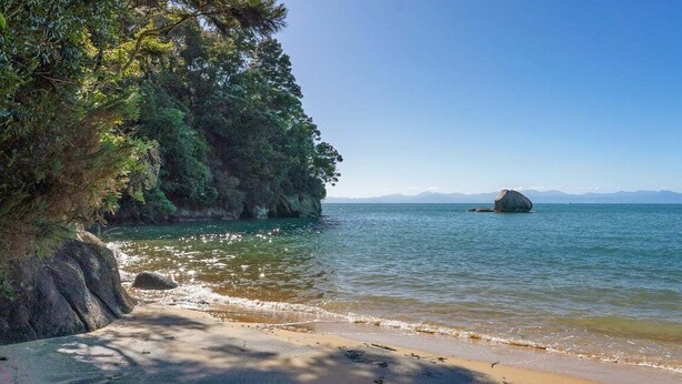 A beach with clear water, a large rock formation, and a mountain range in the distance. - Innisfree Marahau Retreat (Kaiteriteri - Mārahau)