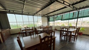 A spacious dining area with multiple wooden tables and chairs, large windows offering a view of the cityscape, and a high ceiling with metal beams. - Hotel Kashi Inn (Jodhpur)