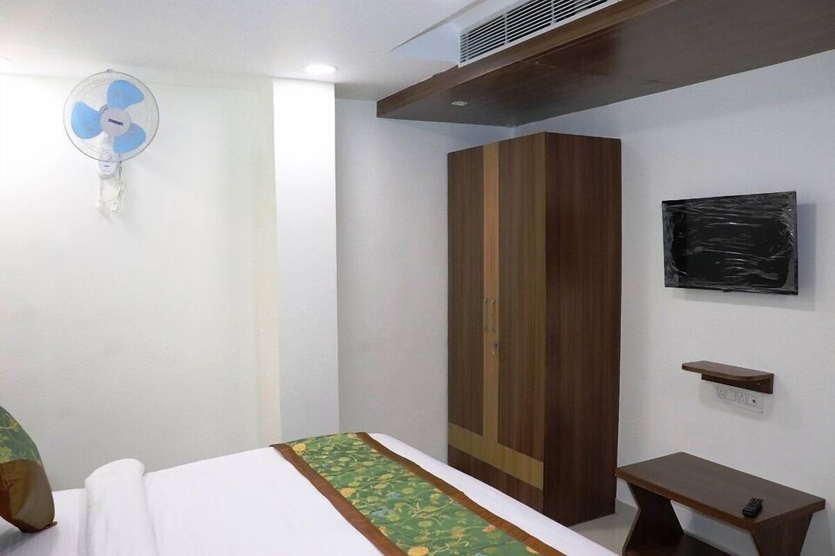A bedroom with a bed, a wardrobe, a bedside table, a wall-mounted fan, and a small shelf.