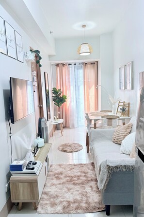 Una sala moderna con sofá, mesa de centro y televisor. Hay un comedor con mesa y sillas. La habitación tiene un ventanal con cortinas.