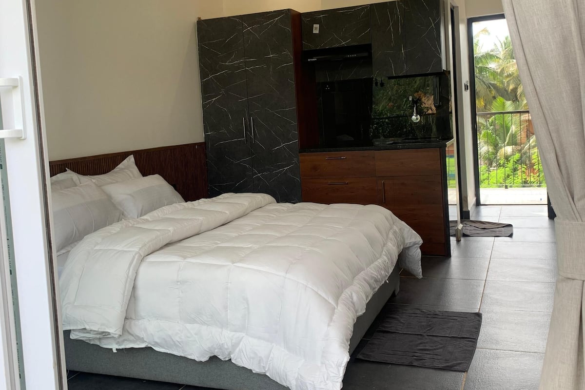 A bedroom with a bed, a wooden wardrobe, a black marble cabinet, and a view of the outdoors.