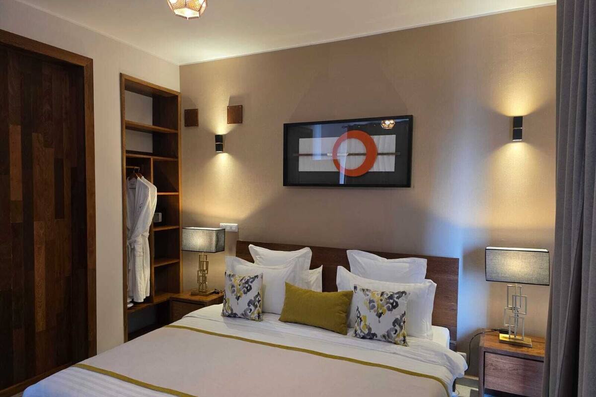 Een hotelkamer met een groot bed, een kledingkast, een nachtkastje met een lamp en een aan de muur hangend kunstwerk.