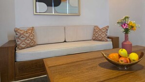 Uma sala de estar moderna com um sofá, almofadas estampadas, uma peça decorativa na parede e uma saladeira com frutas sobre uma mesa de madeira.