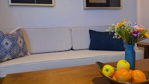 Uma sala de estar com sofá, mesa de centro com frutas, vaso de flores e duas luminárias fixadas na parede.