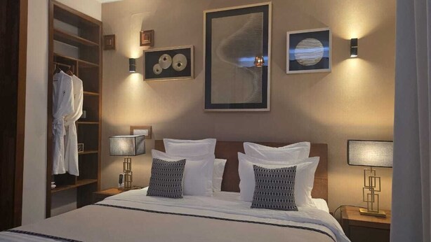 Quarto de hotel com uma cama grande, duas mesas de cabeceira com abajures, um guarda-roupa e obras de arte nas paredes.