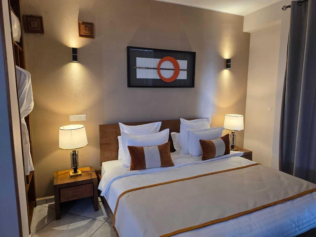 Hotellihuone, jossa on suuri sänky, kaksi yöpöytää valaisimineen, seinälle ripustettu taideteos ja verho oikealla puolella.