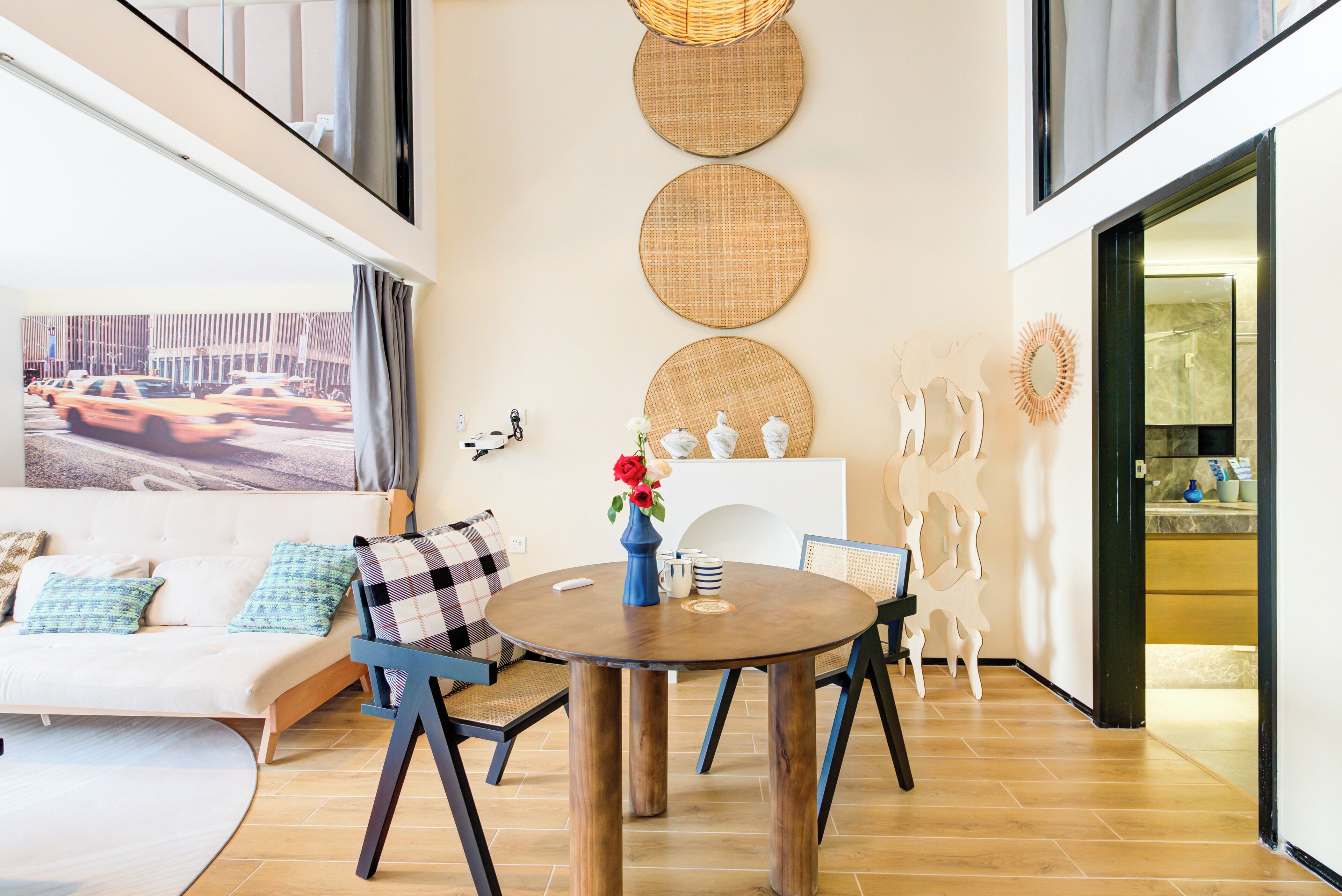 A modern living room with a round wooden table, blue chairs, a white sofa with patterned cushions, and a large window with a cityscape view.