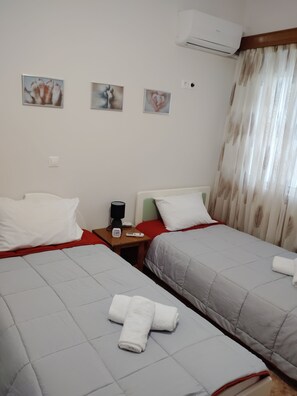 A hotel room with two beds, a nightstand with a lamp, a clock, and a digital device. - Marias Cozy getaway in Glyfada (Glifada)
