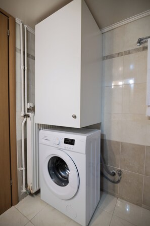 A compact, white washing machine with a control panel, situated in a small laundry room with tiled walls and a wooden door. - Marias Cozy getaway in Glyfada (Glifada)