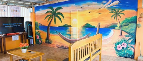 A room with a mural of a beach scene, a wooden bench, a small table with a flower pot, and a TV on a stand.