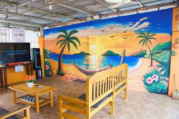 Une pièce décorée d’une fresque représentant une scène de plage, avec un banc en bois, une petite table sur laquelle est posé un pot de fleurs et un téléviseur sur un meuble.