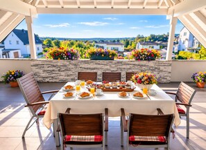 Un espacio comedor al aire libre cubierto, con una mesa dispuesta para el desayuno, rodeada de sillas y flores en macetas.