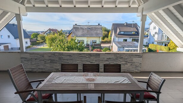 Un patio techado con una mesa y sillas, pared de piedra y vista a casas y follaje.