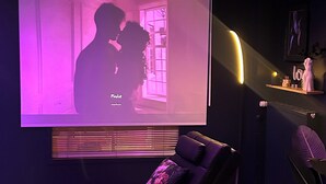 A cozy room with a curved black sofa, a small black table with a candle, a projector screen displaying a silhouette of two people, and a shelf with decorative items. - Romantic gîte, chic love room: L'alcôve Clisson in the heart of the medieval town (Clisson)