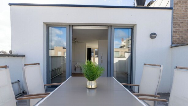 Un balcón con una mesa y sillas, una planta en maceta, y un ventanal corredero que da a un espacio interior.