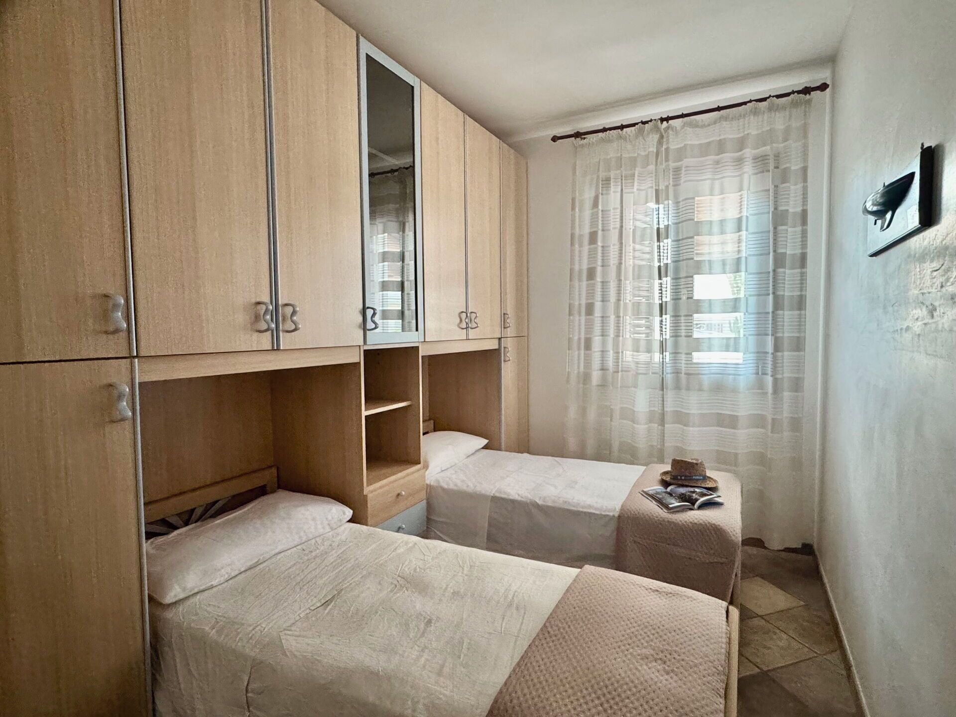 A small, compact room with two single beds, a built-in wardrobe, and a large window with sheer curtains.