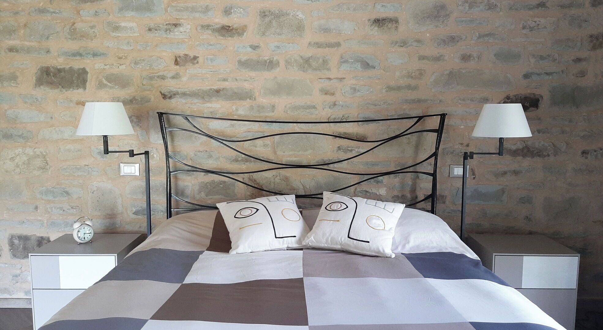 A bed with a checkered bedspread, two bedside lamps, and a clock on the nightstand.