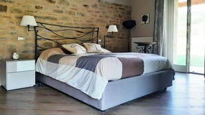 Quarto com parede de pedra, cama com cabeceira cinza, luminárias laterais, mesa de cabeceira com relógio e vista para o exterior através de uma porta de vidro.