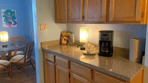 A kitchen with wooden cabinets, a granite countertop, a coffee maker, and a dining area with chairs and a table. - Mermaid's and Mutts Retreat - 30 Day Listing (Norfolk)